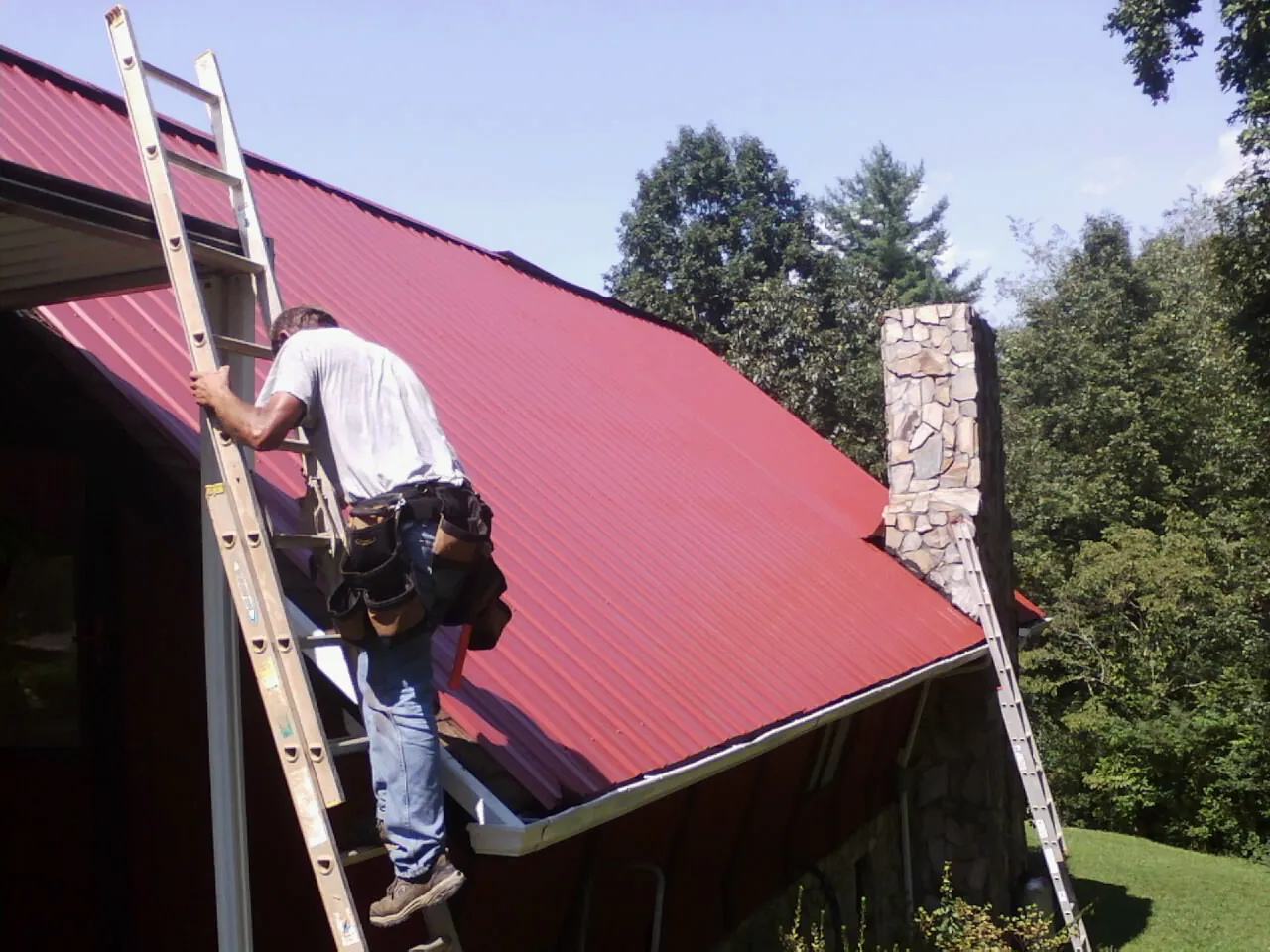 Roofer on a ladder beside a red metal roof and stone chimney — Soffit & Fascia Repair by Roofing Brothers in Alburnett